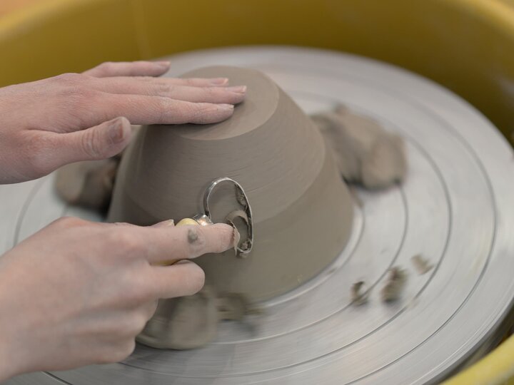 Hands carve at a lump of clay spinning on a potter's wheel.