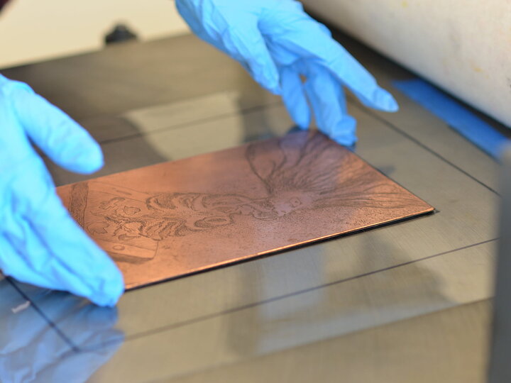 Blue gloved hands adjust a copper etching plate.