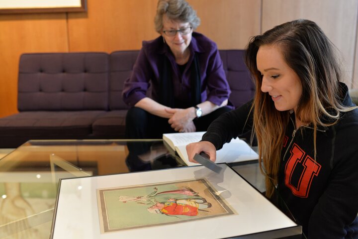 A student studies a book with a magnifying glass as a professor watches with interest.
