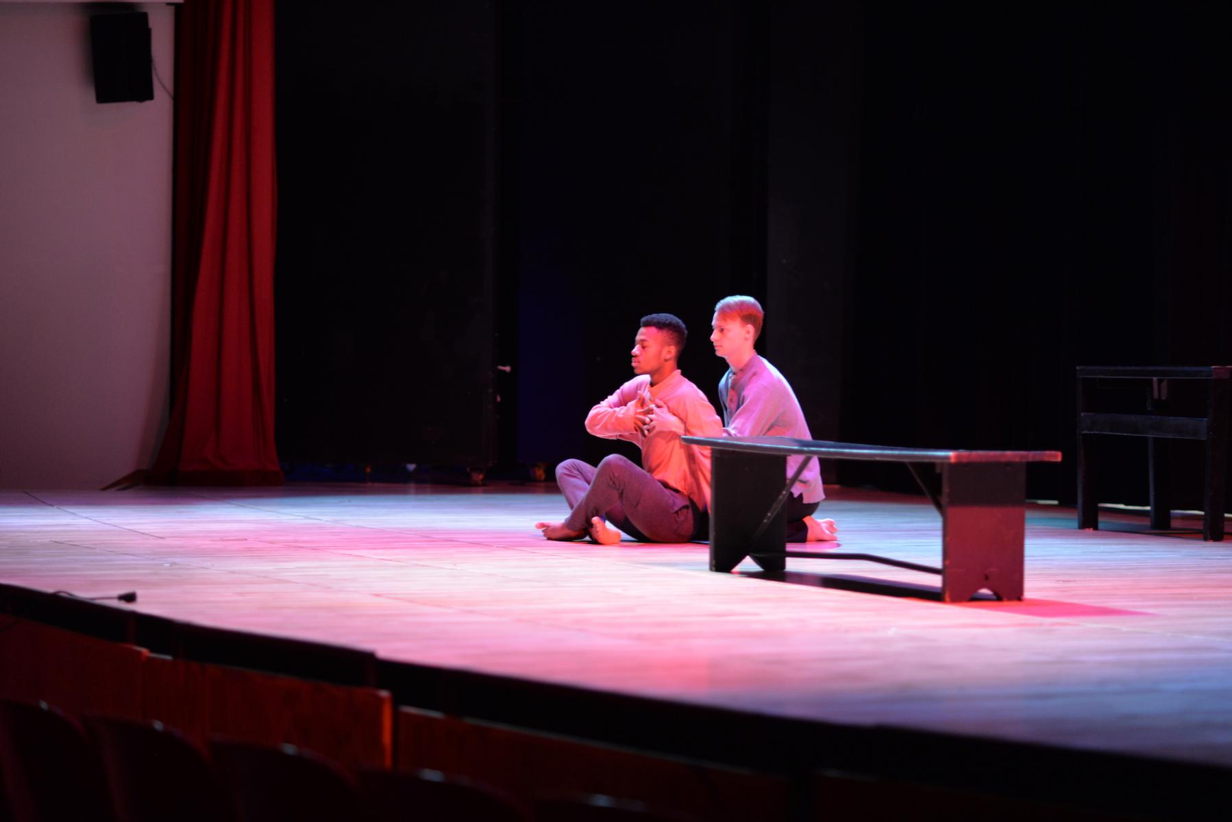 Dance piece performance image, two men sitting together on the ground holding hands.