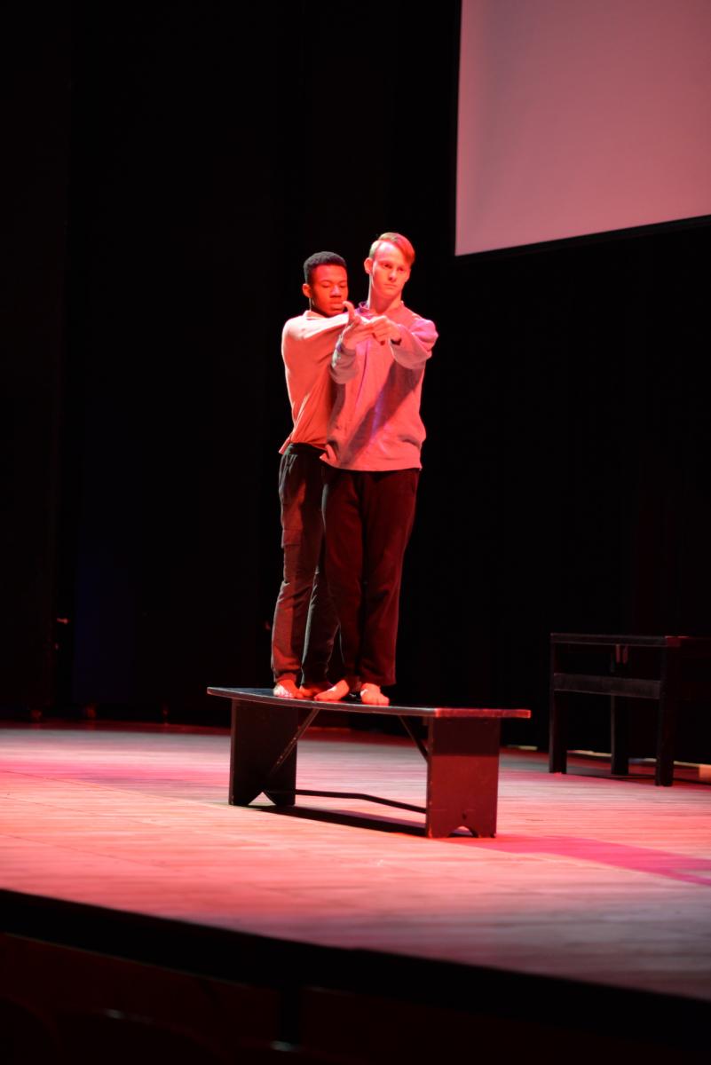 Dance piece performance image, two men standing on a bench together reaching out in front of one another