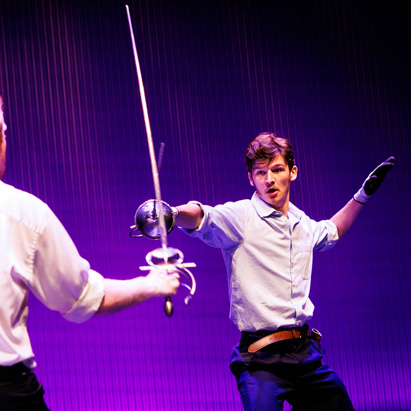 Two individuals fencing under purple stage lighting.