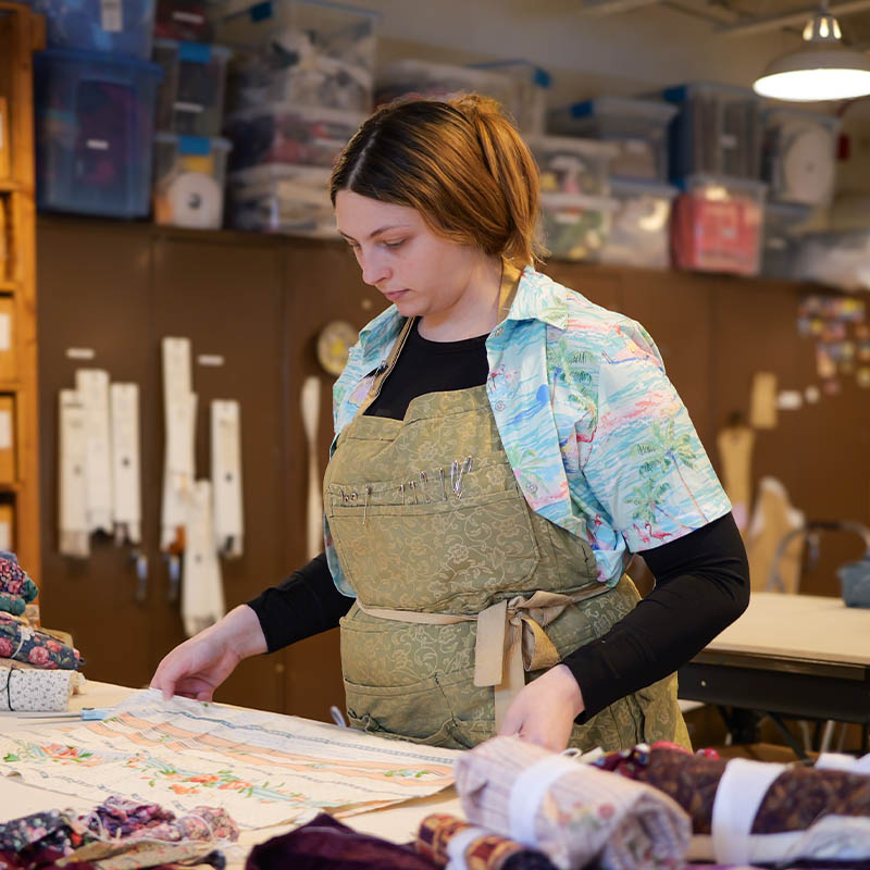 Person in apron organizing fabric on a table in a craft room with shelves and storage boxes.