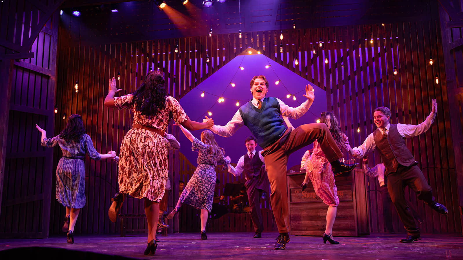 Dancers perform energetically under warm stage lights, wearing vintage-style outfits on a colorful set.