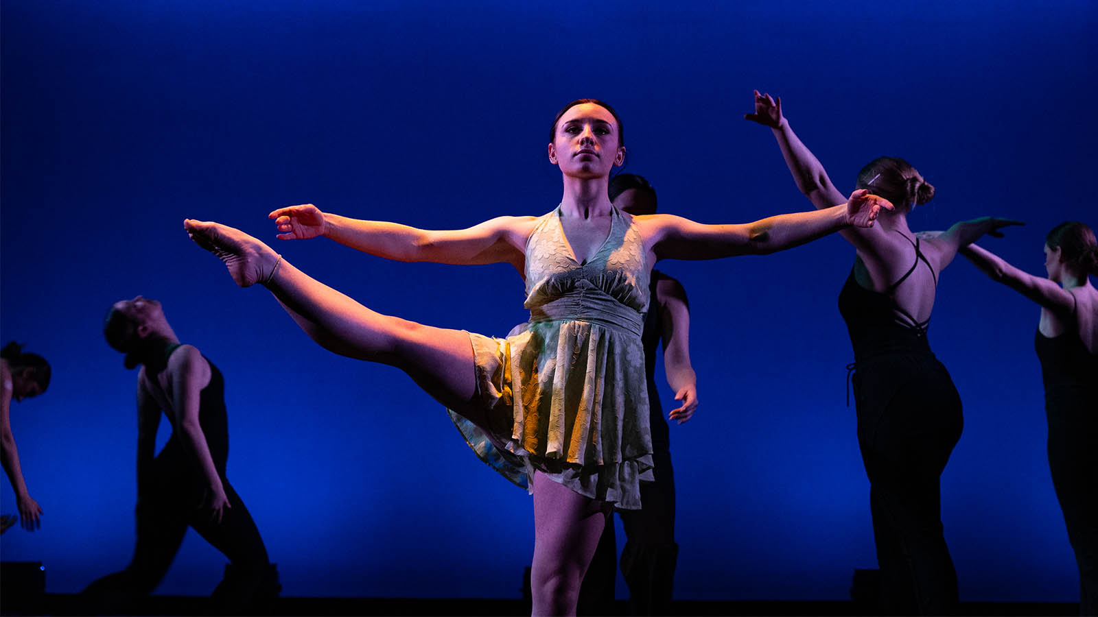 Dancer performs on stage with graceful pose, surrounded by others against a blue-lit background.