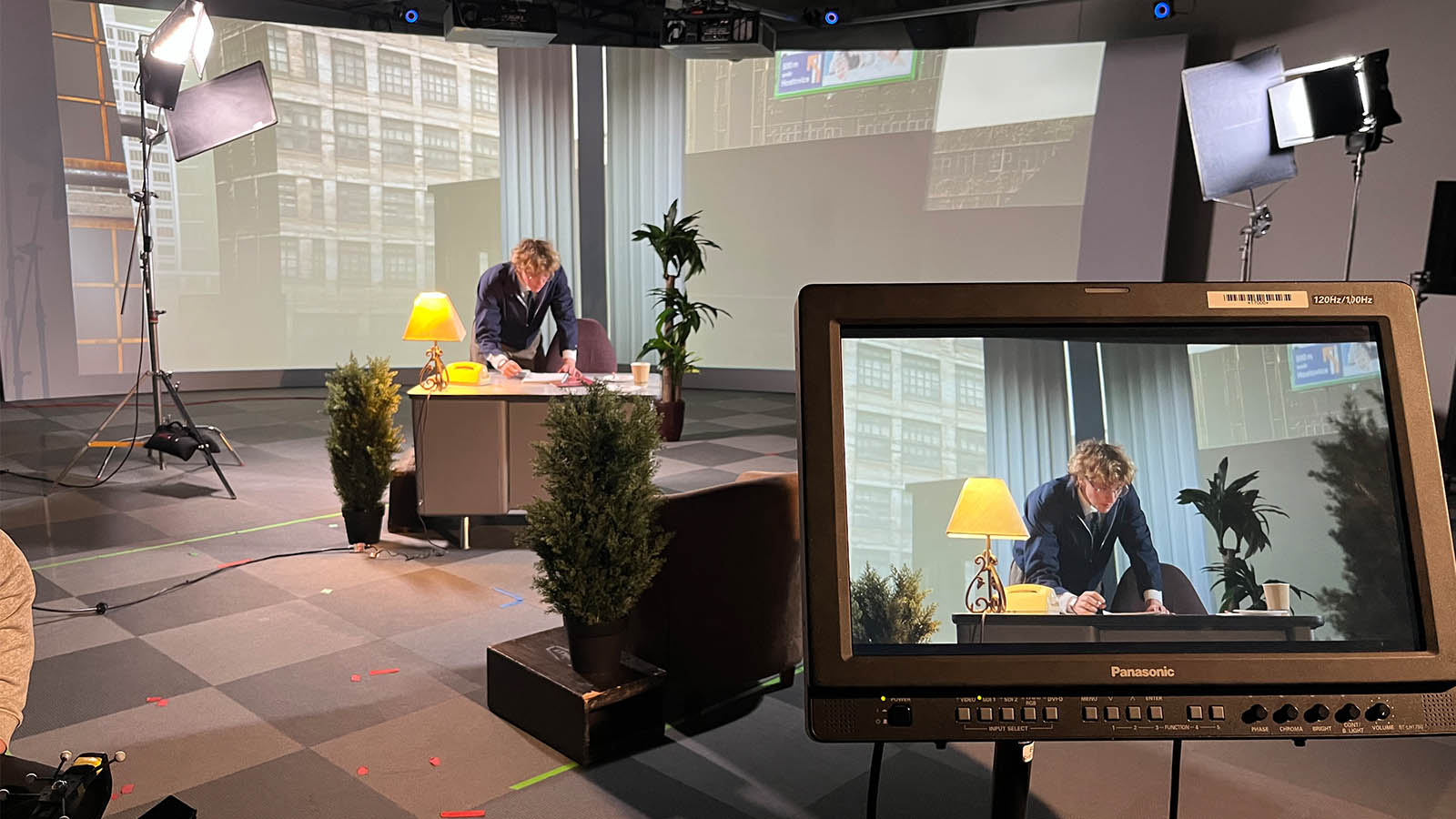 A man works at a desk in a studio with lights and a monitor displaying the same scene.