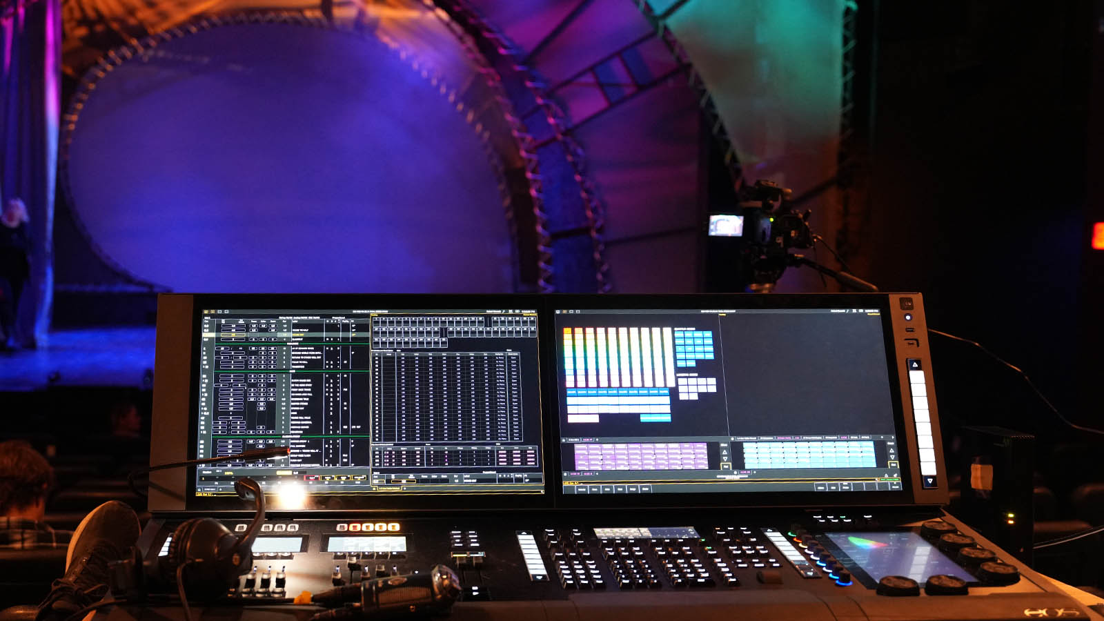 Control desk with lit screens and knobs in a theater; colorful stage lighting in the background.