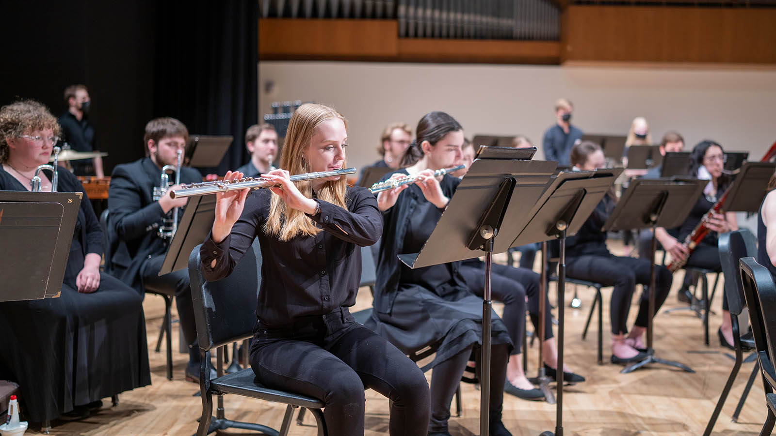 Musicians seated in rows play flutes and other instruments on a concert stage.