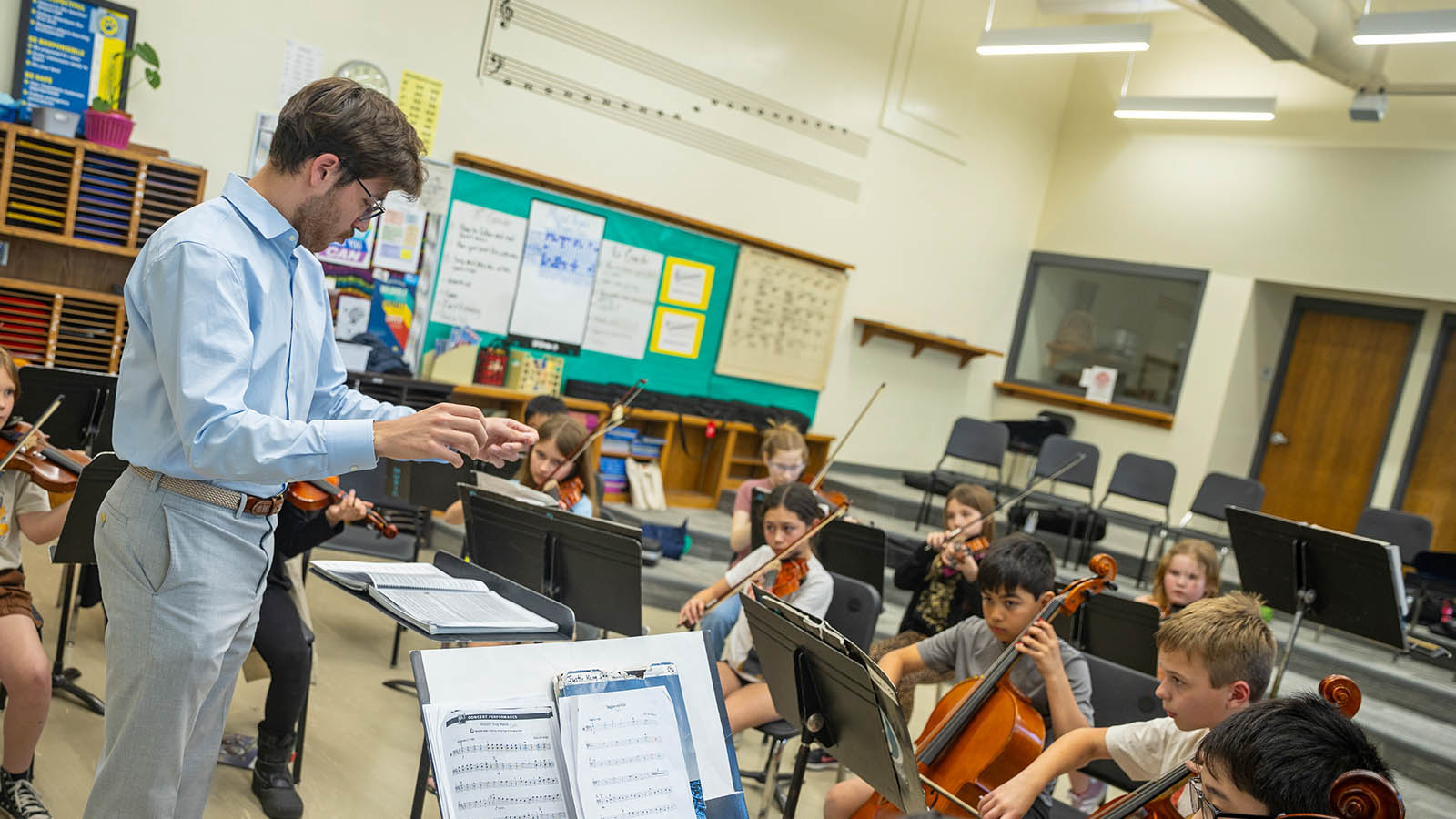 A teacher conducting a group of students playing string instruments in a classroom setup.