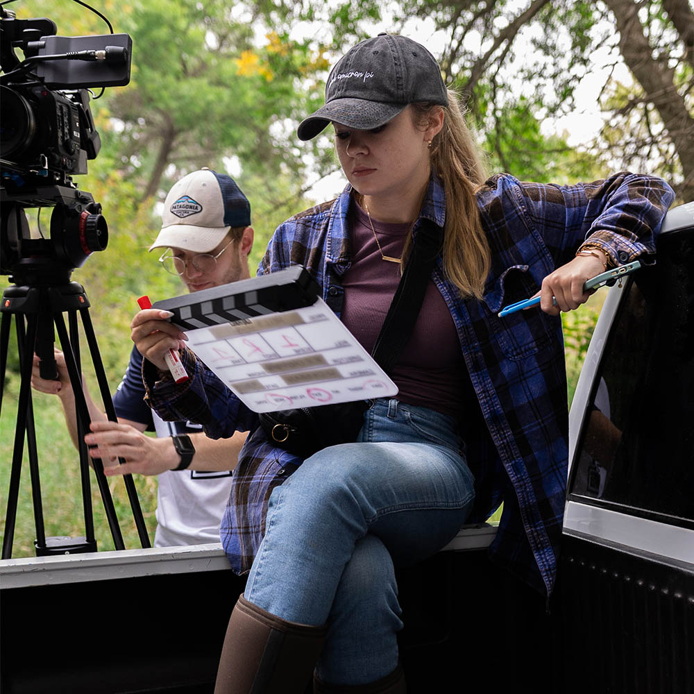 Crew member holding a clapperboard, seated near a camera setup outdoors with greenery in the background.