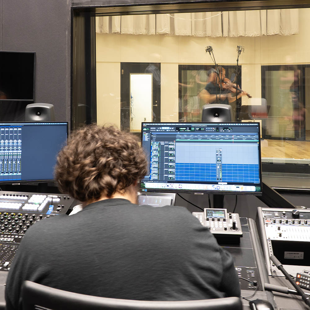 Recording studio with sound engineer at mixing desk, glass window showing musician playing violin.