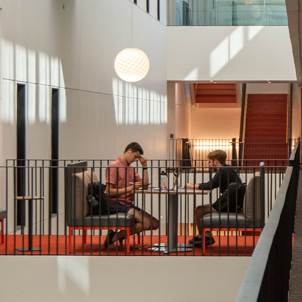 Two people seated at a table in a modern, sunlit space with red stairs in the background.