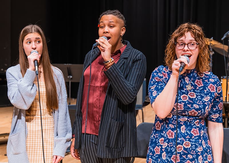 Three singers stand close together on stage singing into microphones.