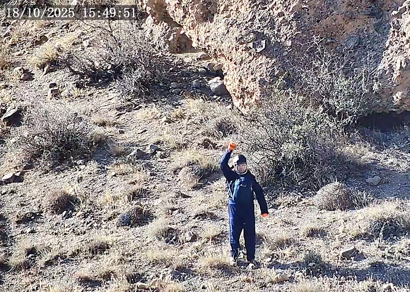 Person in a blue outfit holding an orange object, standing in a rocky, shrub-filled landscape.