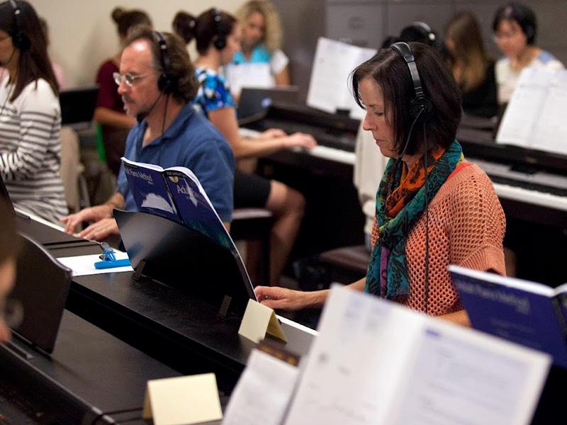 A room full of several people at pianos with headphones on playing the piano.