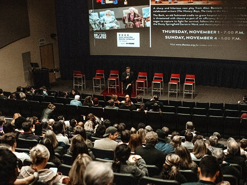 An audience in The Ross theatre at a talk with the speaker in front of the theatre screen