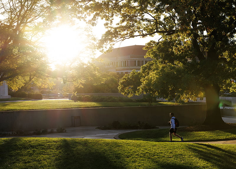 A person walk across campus with early morning sunlight silhouetting them.