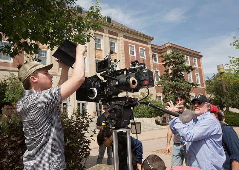 A film camera is adjusted for a shot with guest artist giving direction.