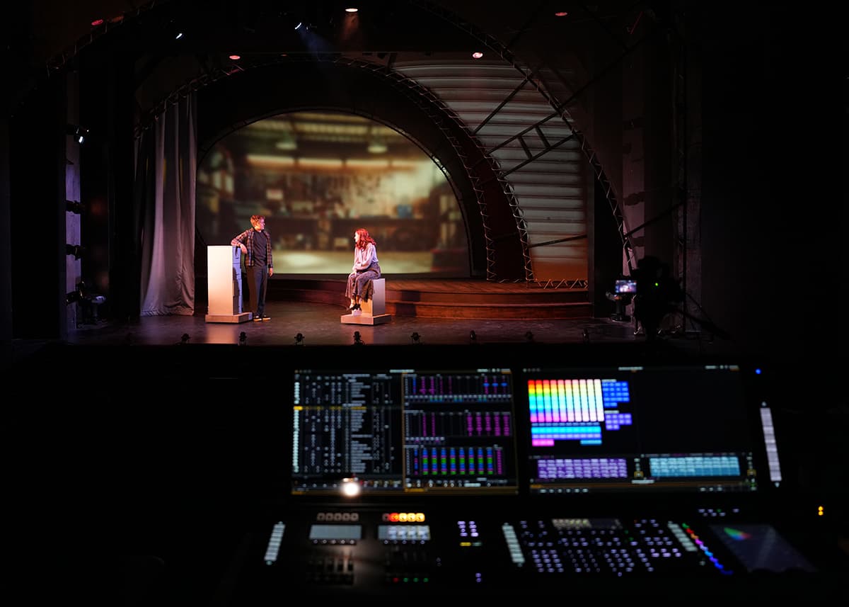 Two actors block out a scene on stage while the theatre control panel sit alight within the darkened theatre.