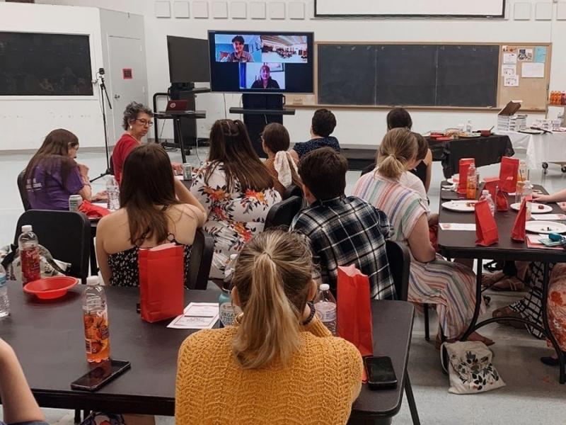 Photo from back of a classroom where members of SAI are sitting at tables watching a video conference on a screen in the front of the room.