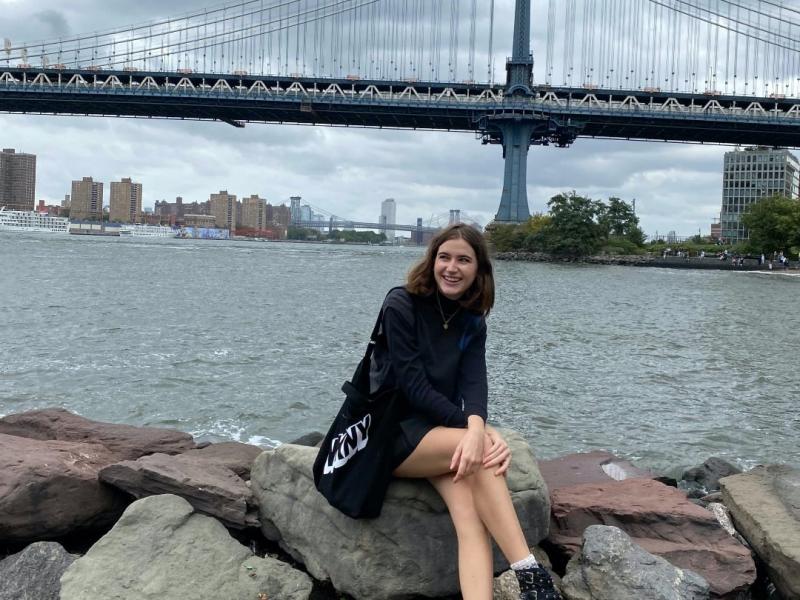 Sabrina Sommer, smiling and looking off-frame while sitting on a large rock in front of a body of water and bridge in New York City.   