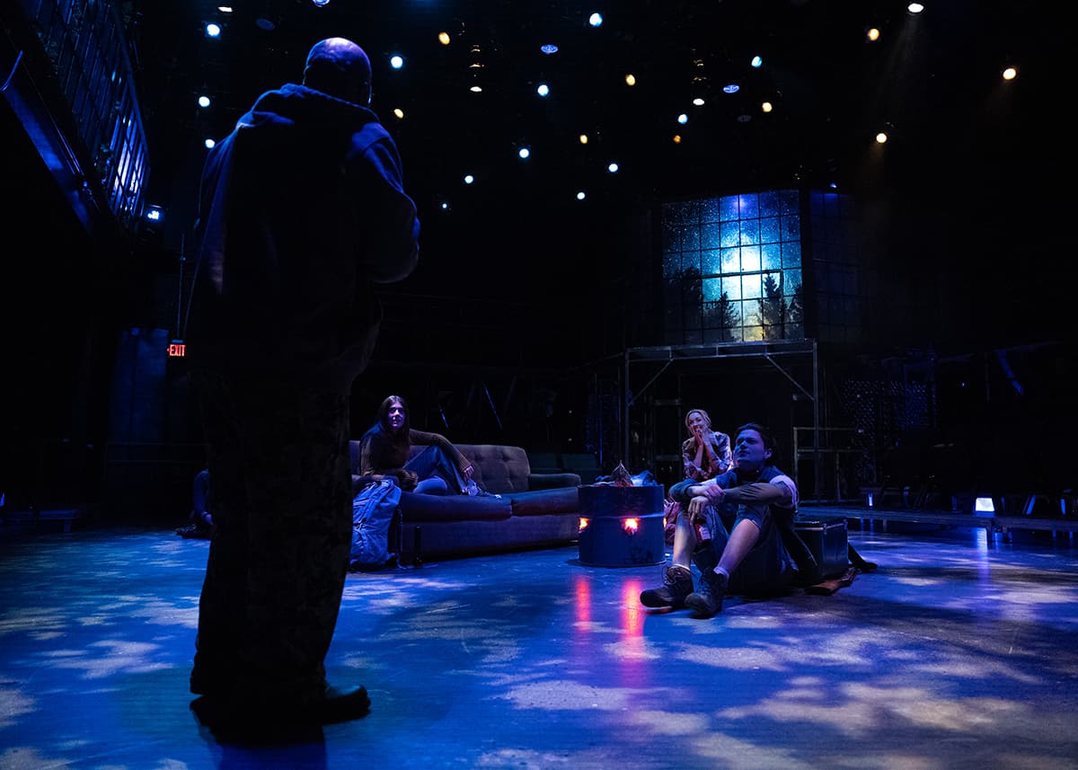 A dark figure stands in the foreground with actors sitting listening on a darkened stage.