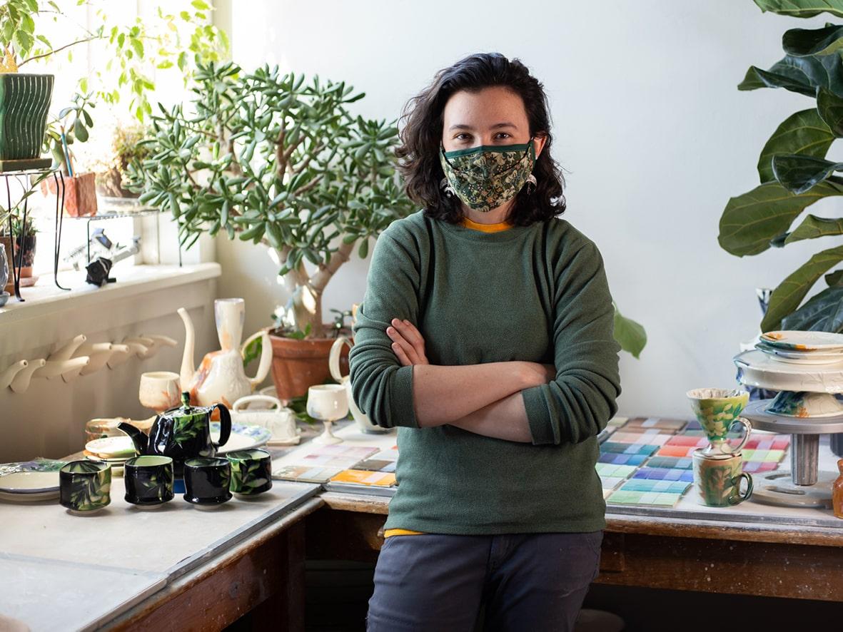 Portrait of a ceramics student in front of working surfaces with plants, ceramic works, and samples of glaze studies in a sunny, art studio.