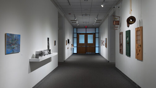 An image of the main gallery with art on both the left and right sides extending down the hall to a set of closed double doors. There are open doorways into the east and west galleries midway through the parallel walls.