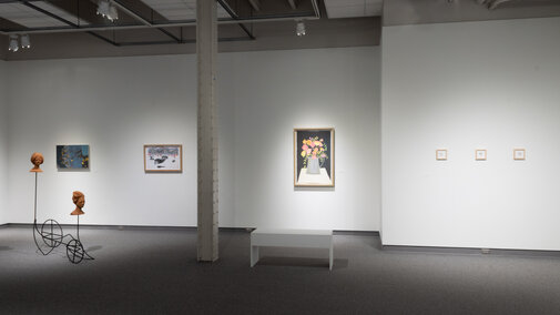 An image of a gallery where there are multiple pieces hanging on the back wall, with a sculpture placed on the ground on the lefthand side and a bench placed directly in front of a large piece on the wall.