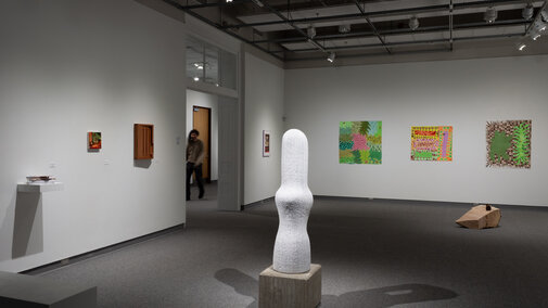 Image of a gallery with a sculpture in the foreground, a shelf with books, a painting, and a wooden cabinet on the left wall, and 3 large prints on the wall in the background with one framed photo on the adjacent wall.