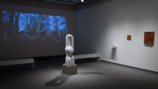 An image of a gallery with a projected video on the back wall on the left side of the frame with two low benches between the wall and a large sculpture in the middle of the room. There is a shelf on the right wall with a small painting and wooden cabinet to the right on the same wall.