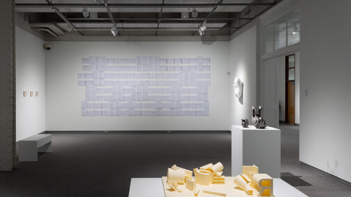 image of a gallery with sculpture on a pedestal in the foreground, a sculpture on the right on a pedestal, installation of art on the background far wall, and other art hanging at eye level on the left and ride sides of the gallery walls