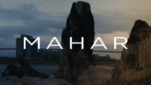Text reads "MAHAR" with an image behind of two large tree stumps in a riverside setting at dusk with a bridge visible in the background.