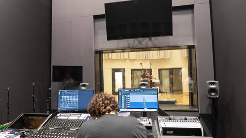 Audio engineer working at a control panel in a recording studio, viewing musicians through a window.