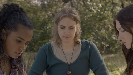 Still from seven doors. Three people sitting outdoors in a wooded area, faces blurred; sunlight filtering through trees.