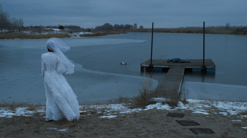 Still image from the film of a Woman in a white dress by a frozen lake, with a dock and figure lying motionless in the distance.