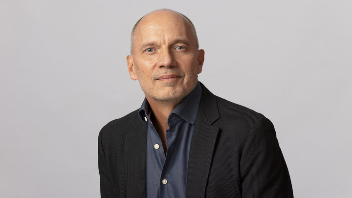 Paul Barnes in dark blazer and blue shirt against a plain, light gray background.