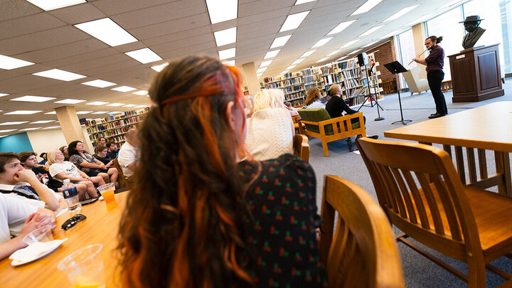 Audience seated in a library, watching a musician play a brass instrument near bookshelves.