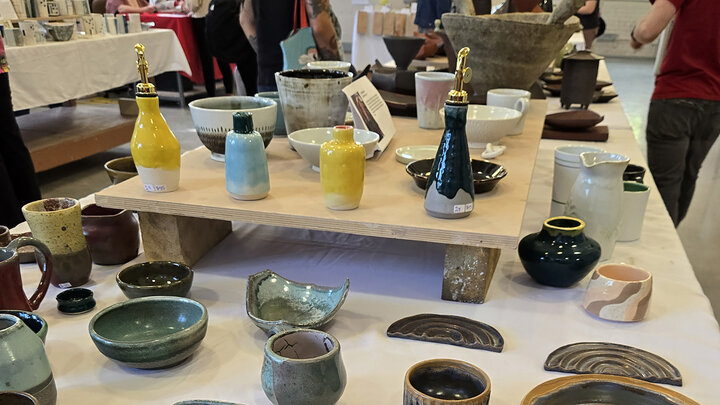 Ceramic display with bowls, cups, and bottles on tables at a market, people browsing in the background.