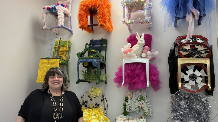 Wall-mounted decorative chairs with vibrant designs; The artist Kimberley D'Adamo smiling, dressed in black; studio setting.