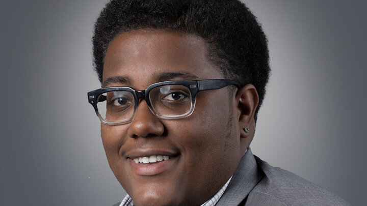 Headshot of Justin Payne wearing glasses and a gray blazer, facing partially to the side against a neutral background.