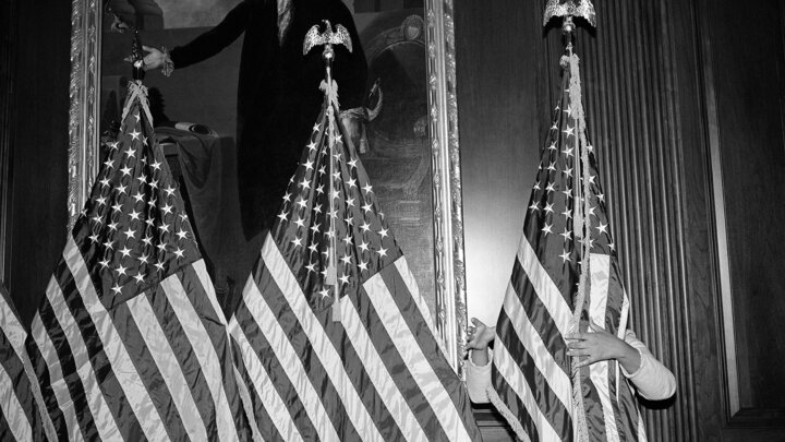 An aide to House Speaker Nancy Pelosi adjusts a row of flags in front of a painting of George Washington at the U.S. Capitol, preparing for Democratic leaders to speak after the vote on December 18, 2019, to impeach President Donald Trump the first time. The House impeached the president for abuse of power and obstruction of Congress for trying to get Ukraine to investigate Joe Biden. Â© Louie Palu. 