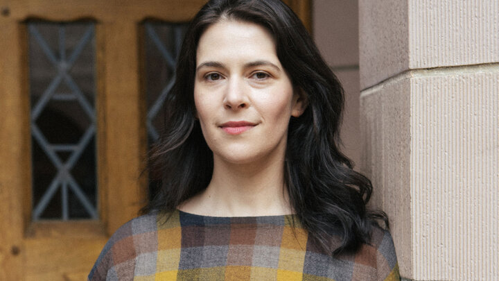 Juliet Sperling with dark hair in a plaid top, standing near a wooden door and stone wall.