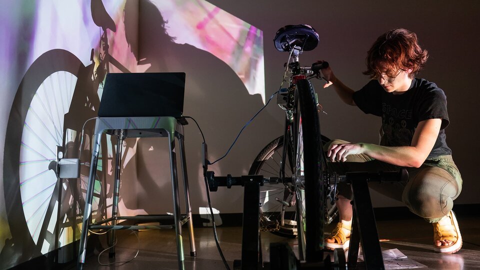 A student kneels next to a bike, hooked to a laptop, pedaling it by hand and illuminated by a projected image.