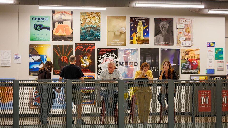 Visitors to the open studios stand at a railing with a wall covered in project posters behind them.