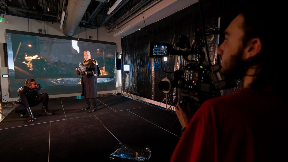 Two actors in sci-fi costumes stand in the background playing out a scene on a virtual set, a camera person filming in the foreground.