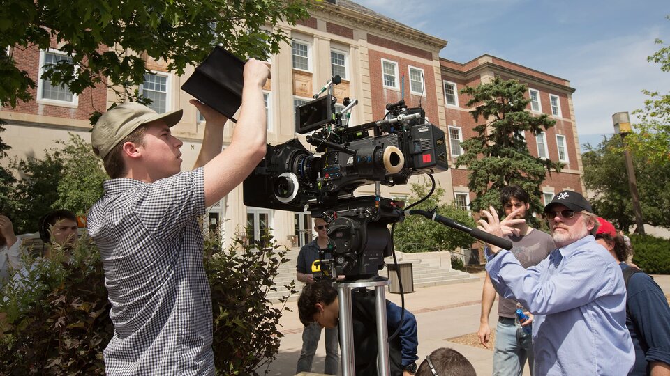 A film camera is adjusted for a shot with guest artist giving direction.