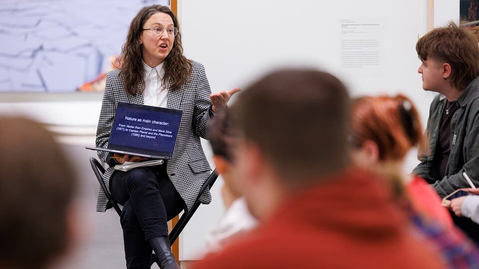 A professor lectures to students while holding a laptop up for them to see.