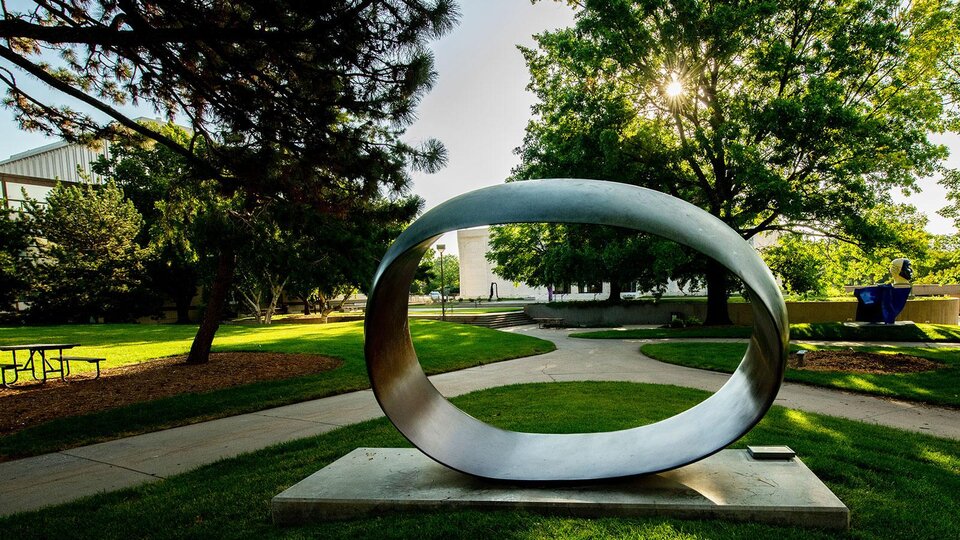 large outdoor sculpture in an oval shape with the sunrise shining through green trees