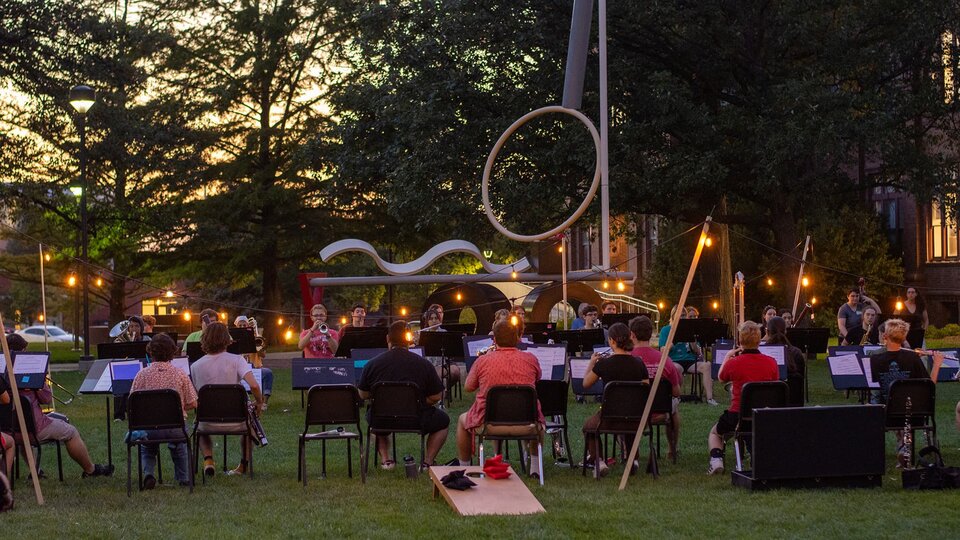 An music ensemble plays outside on the greenspace with a large sculpture in the background.